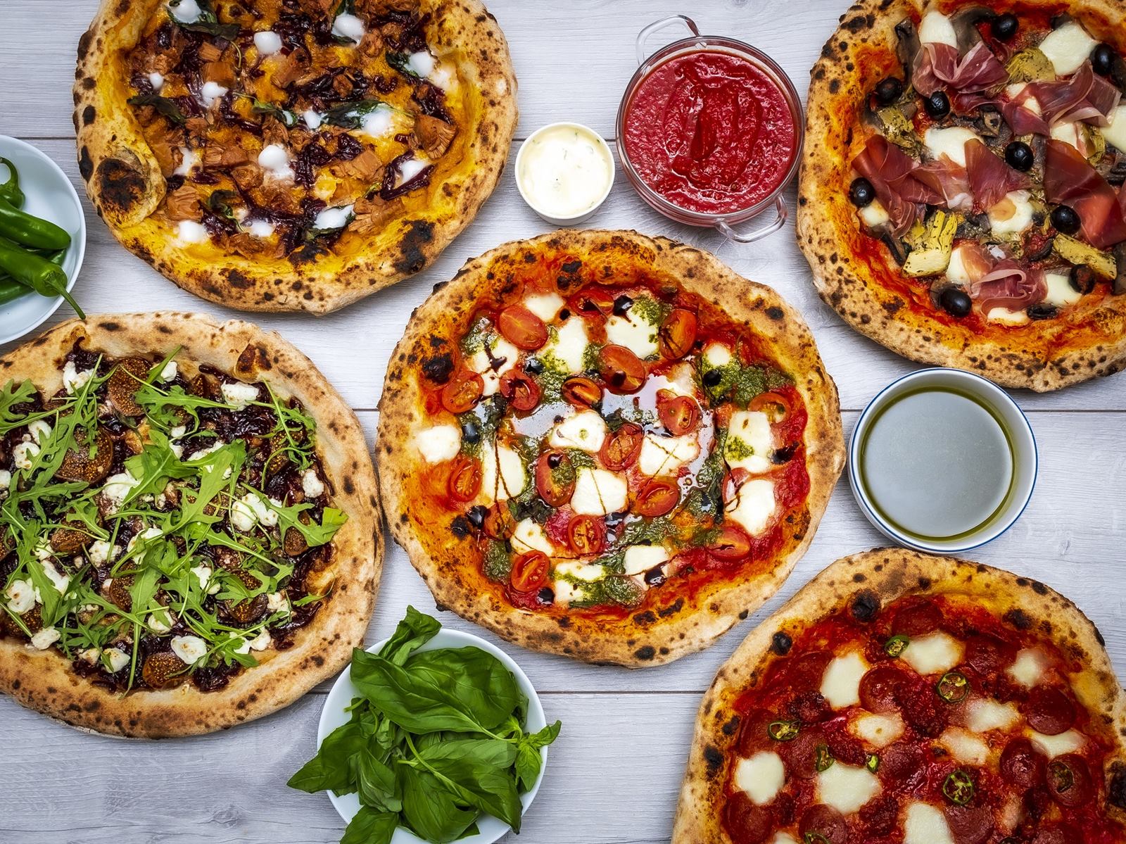 A table spread of Pesto Kitchen pizzas — Caprese, Capricciosa, Figgi Smalls and a spicy pepperoni — with bowls of basil, green chillies, olive oil and tomato sauce